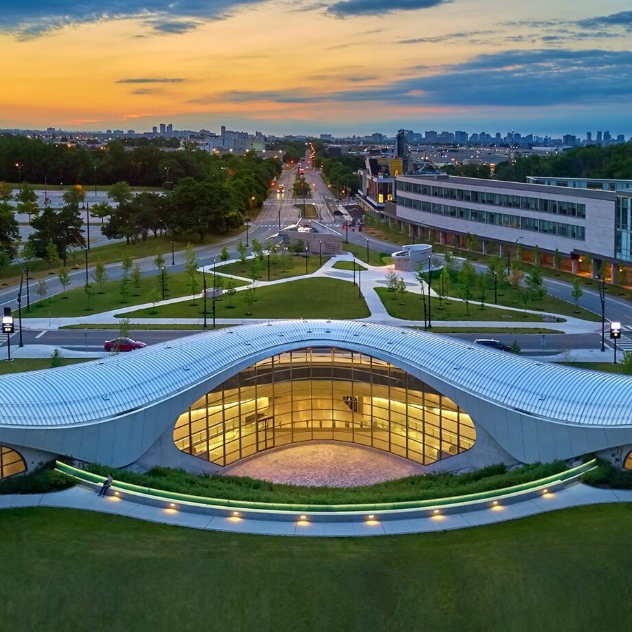 Curved modern building, York University subway station, with large glass windows at dusk with illuminated interior and urban landscape in the background.