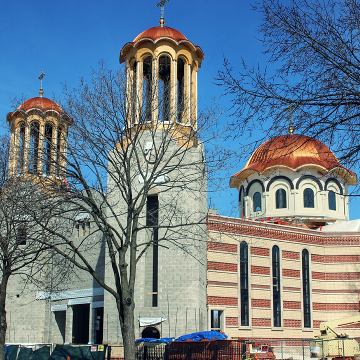 Church building with three domes topped with crosses, one dome has a terracotta roof and arched windows, leafless trees in front under a clear blue sky.