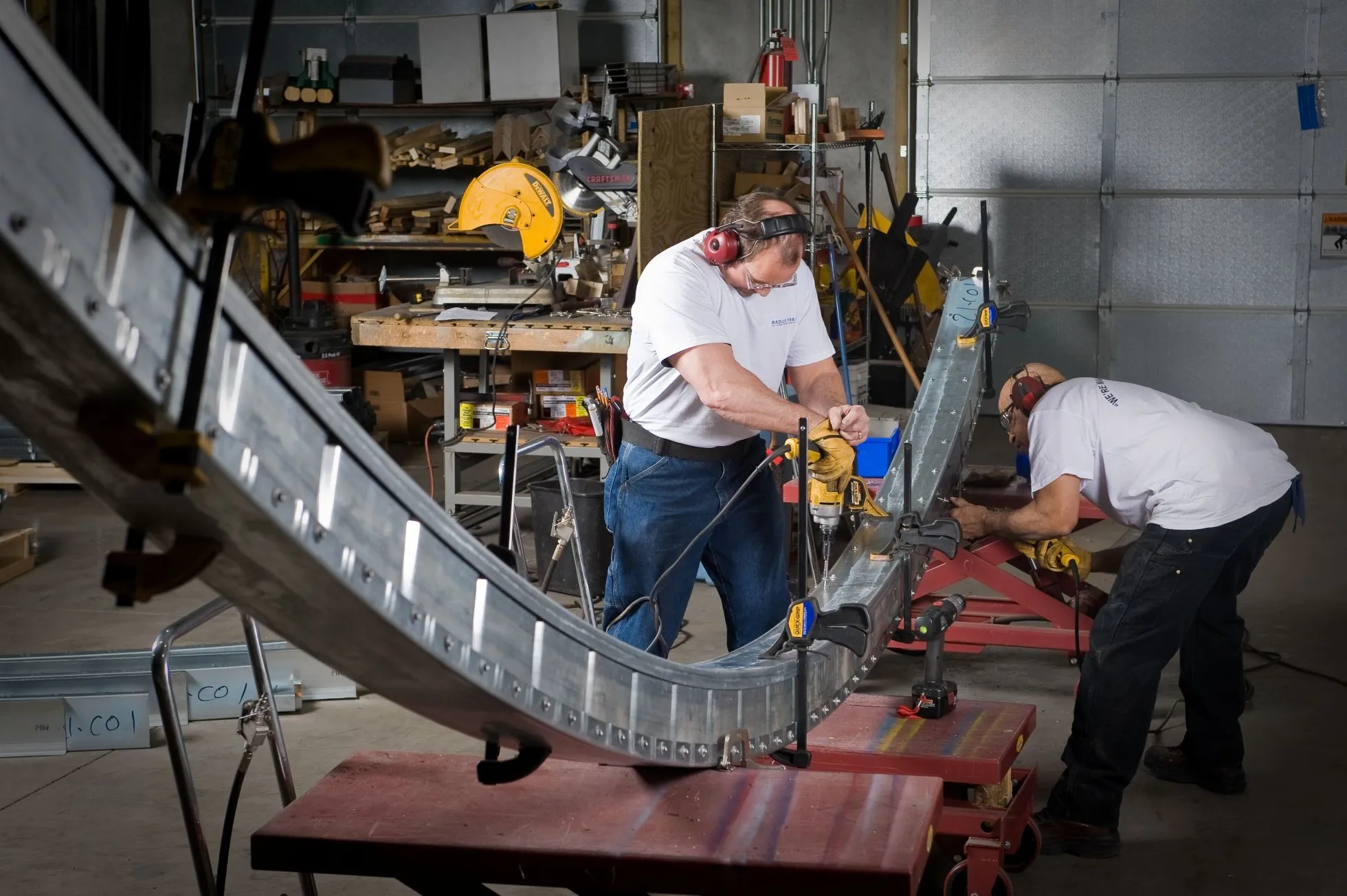 Two workers in a fabrication workshop assembling a large curved metal box beam using clamps and power tools.