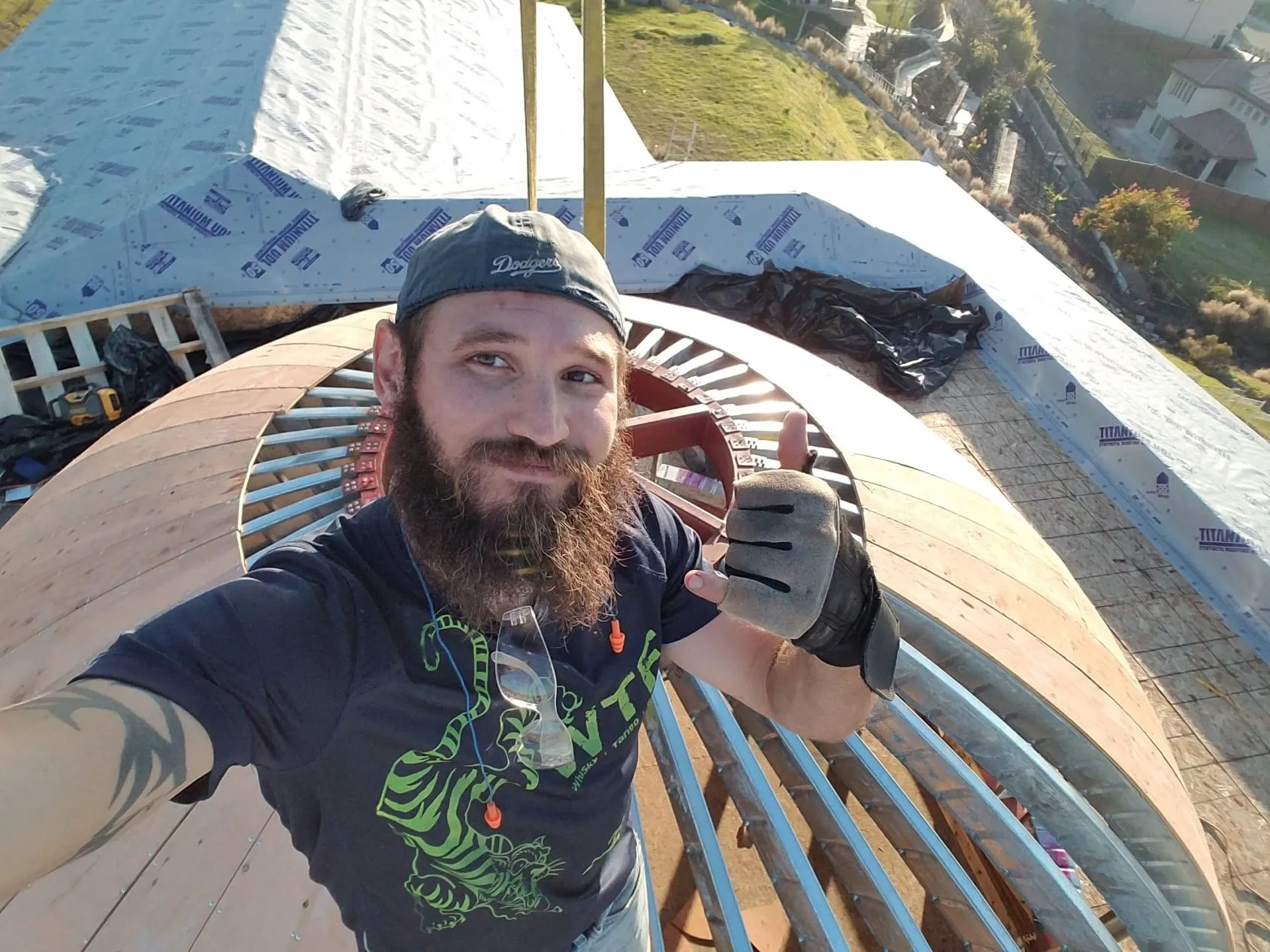 Bearded man wearing a backwards Dodgers cap and a black glove taking a selfie on a dome rooftop under construction giving a hang loose hand signal