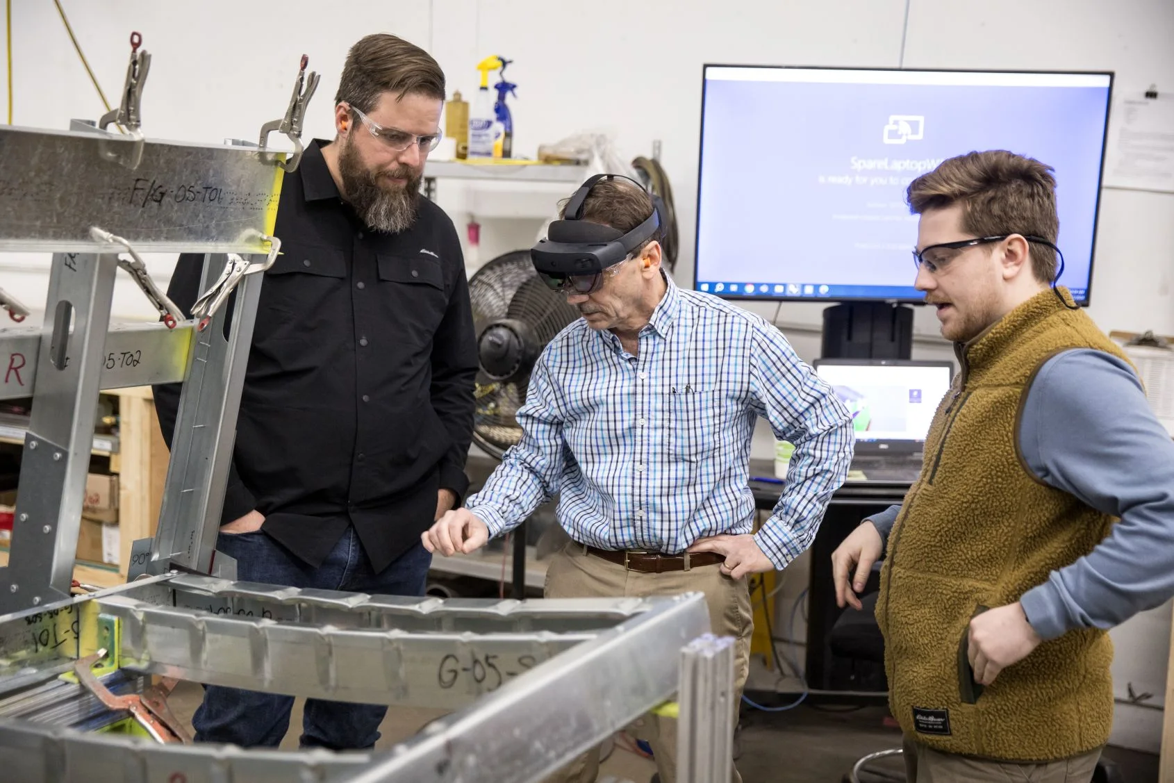 Three men wearing safety glasses examine a metal framing with one man using augmented reality headset in a workshop.