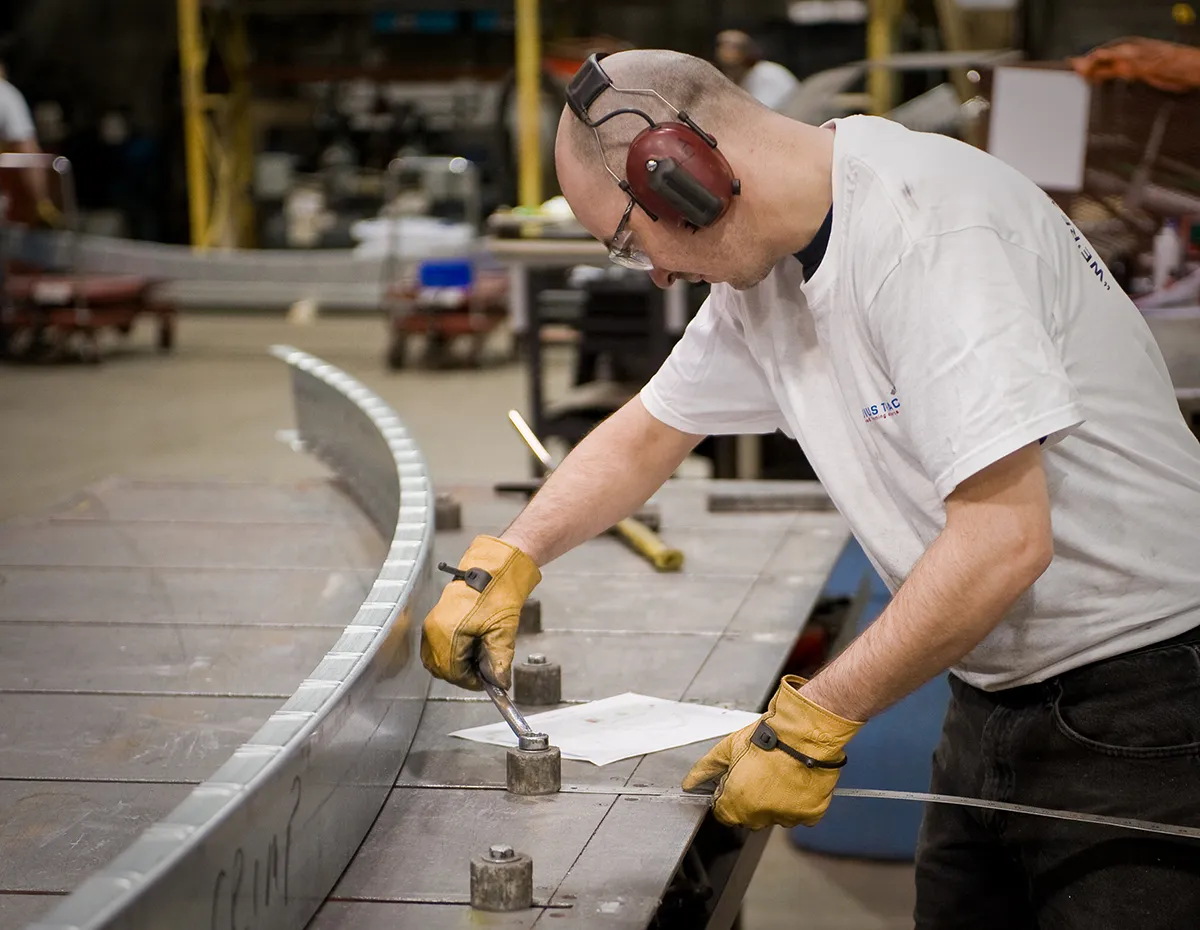 Worker in safety gloves and ear protection prepares for a quality assurance check and adjusts a metal track segment in an industrial workshop.