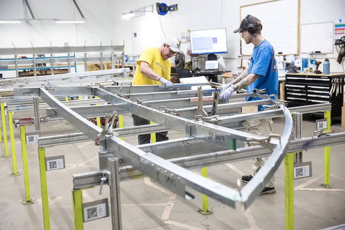 Two workers assembling a large metal framing panel on yellow jig stands in a workshop.