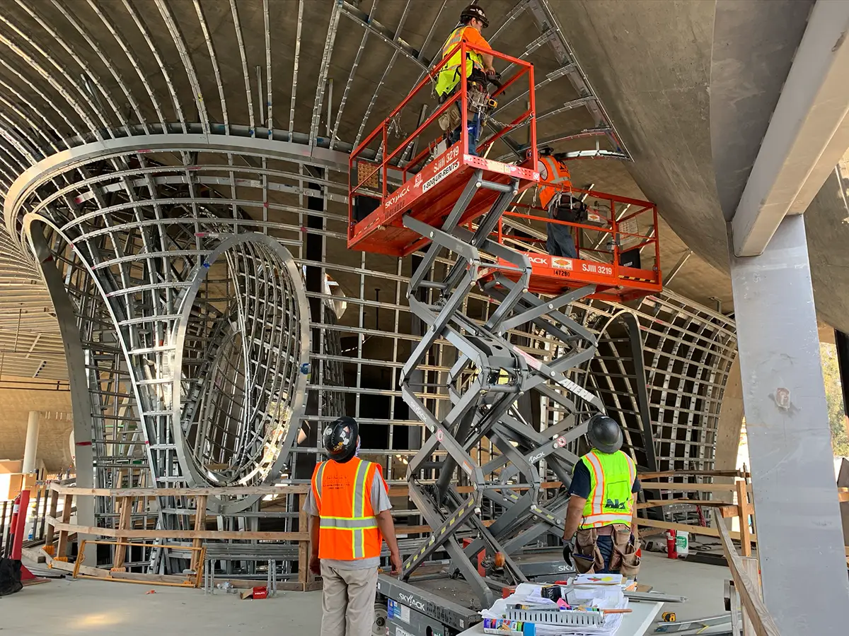 Construction workers installing large curved panelized metal framing under a concrete ceiling using a scissor lift.