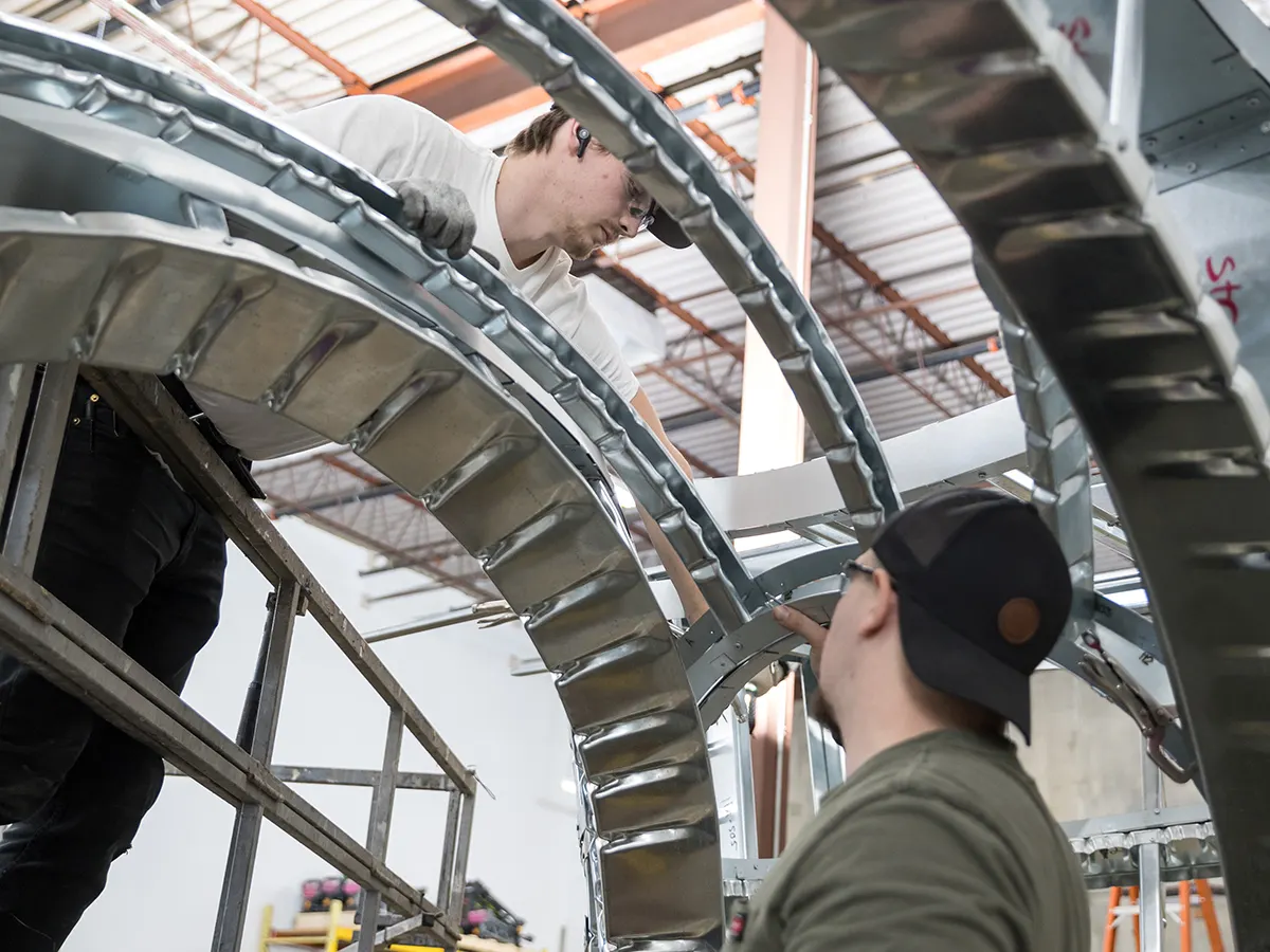 Two workers assembling curved metal framing panels inside a warehouse.