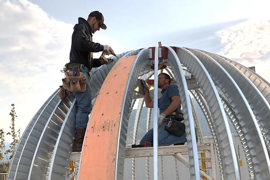 Two construction workers assembling a metal dome frame, with one installing laser cut to size plywood.