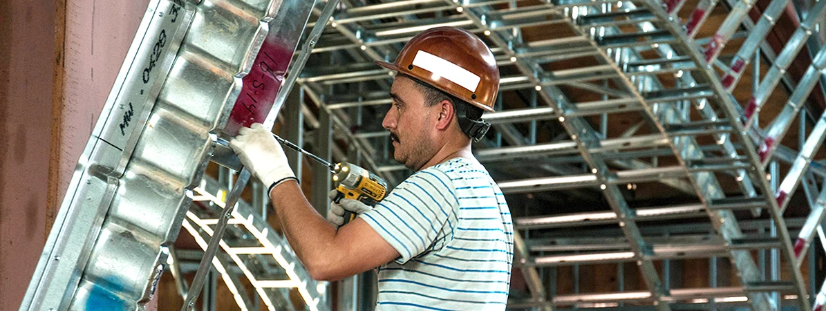 Construction worker wearing a brown hard hat and gloves uses a drill on metal framework inside a building under construction.