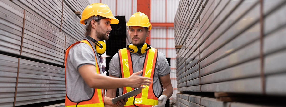 Two logistic workers wearing yellow helmets and orange safety vests discussing with one holding a tablet in a warehouse with stacked metal framing.