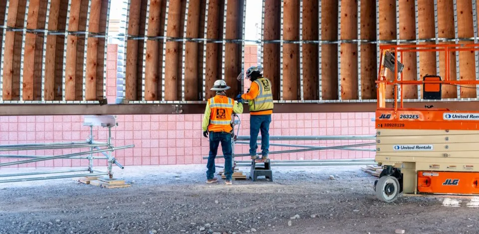 Two construction workers in safety vests and helmets standing inside a large metal framed dome structure under construction.