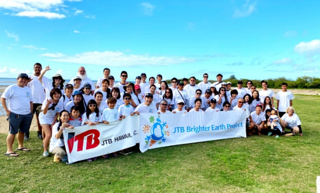 A large group of volunteers participating in the JTB Brighter Earth Project in Hawaii, gathering outdoors under a bright blue sky to promote environmental awareness.