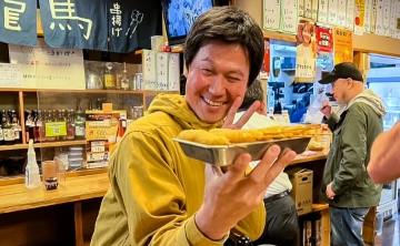 Man smiling and posing with a plate of kushikatsu (deep-fried skewers) at a local Japanese restaurant.