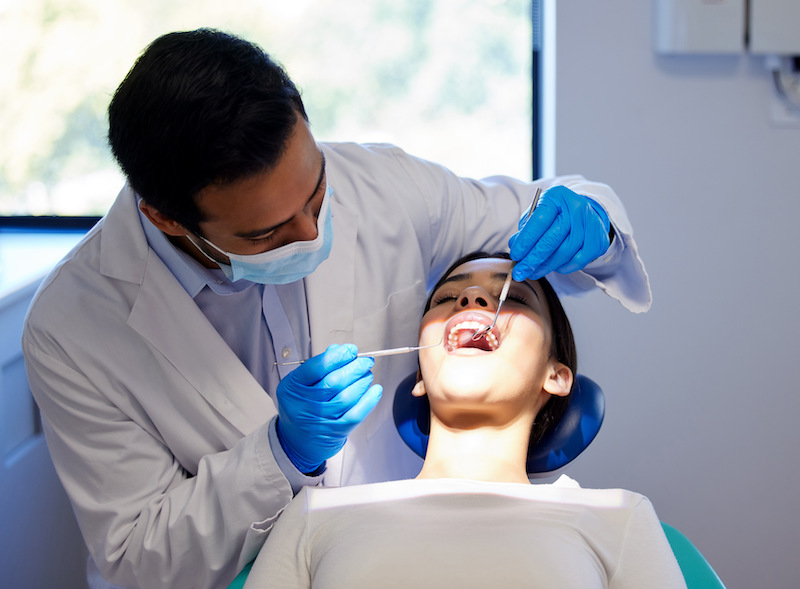 Image showing a dentist performing a dental check-up, which typically takes around 30 minutes to an hour. The dentist is wearing a face mask and blue gloves while examining the patient's teeth, who is reclined in a dental chair.