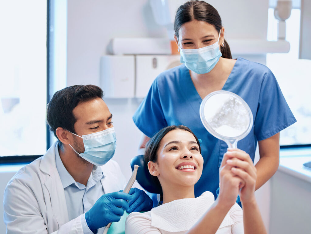 Young woman looking into a mirror and smiling after her dental cleaning, with her dentist and assistant in the background. Private insurance can help you cover dental cleaning costs, keeping your smile even brighter.