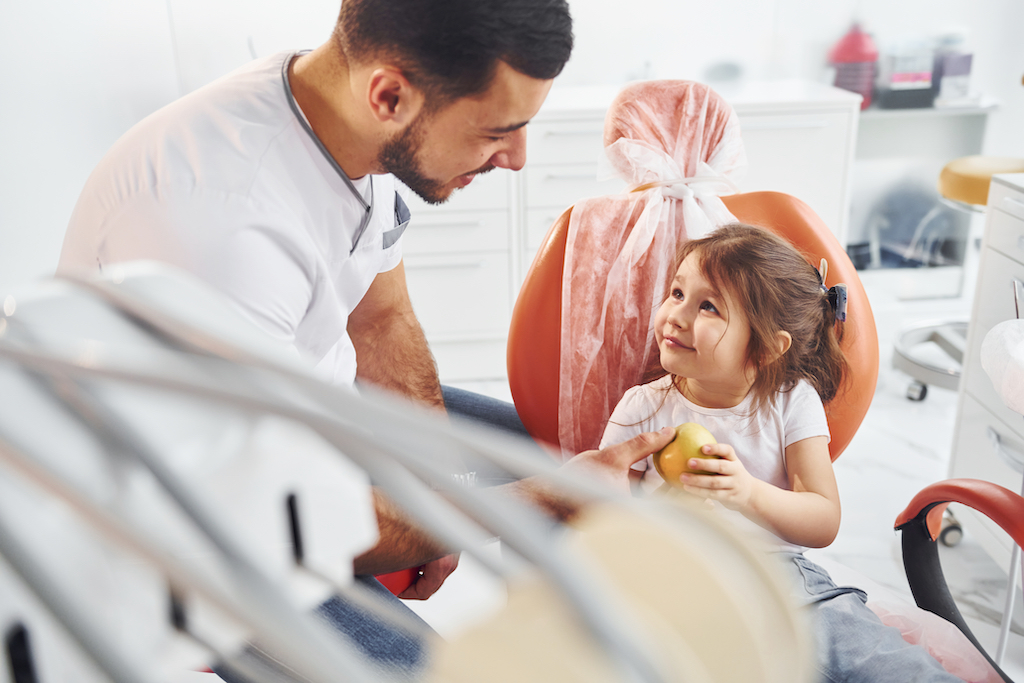 A young girl sits in a dentist's chair holding an apple, highlighting how to prevent cavities in children through regular dental check-ups and healthy eating.