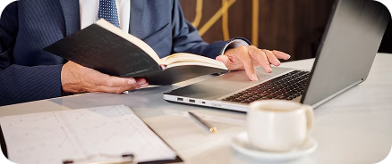 Person in suit reading a book while using a laptop at a desk with papers and a coffee cup.