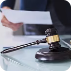 Gavel resting on a sound block on a glass table with a blurred person holding documents in the background.