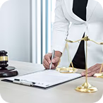 Person signing a document on a clipboard next to a brass balance scale and a wooden gavel on a desk.