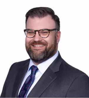 Man with dark hair and beard wearing tortoiseshell glasses, a navy suit, white shirt, and striped tie against a plain background.