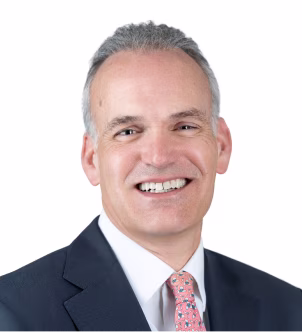 Smiling middle-aged man with short gray hair wearing a dark suit and light pink tie against a dark background.