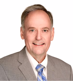 Smiling middle-aged man with short brown hair wearing a dark suit, light blue shirt, and red tie in an office setting.