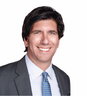 Smiling man with dark hair wearing a gray suit, white shirt, and blue patterned tie against a white background.