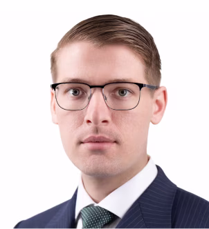 Portrait of a young man with light skin and slicked-back brown hair wearing glasses, a navy suit, white shirt, and green tie.