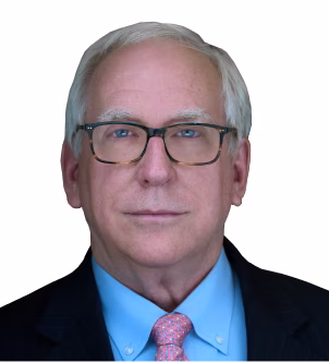 Older man with white hair and glasses wearing a blue shirt, pink tie, and dark blazer on a white background.