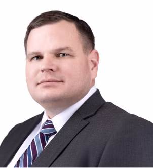 Professional headshot of a man with short dark hair wearing a dark suit, white shirt, and striped tie against a white background.