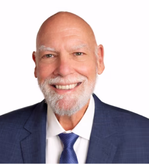 Smiling elderly man with a white beard wearing a dark suit and blue tie on a white background.