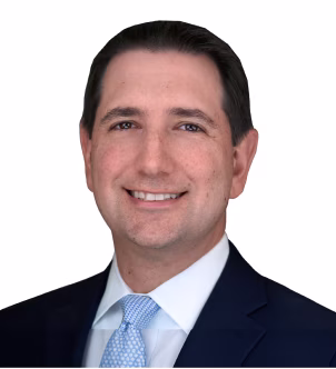 Portrait of a man in a dark suit, white shirt, and red tie smiling against a plain gray background.