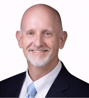 Smiling bald man with a gray beard wearing a dark suit and light blue tie on a white background.