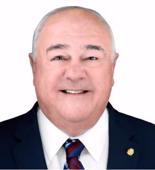 Smiling older man in a dark suit, white shirt, and striped tie with a pin on his lapel, against a white background.