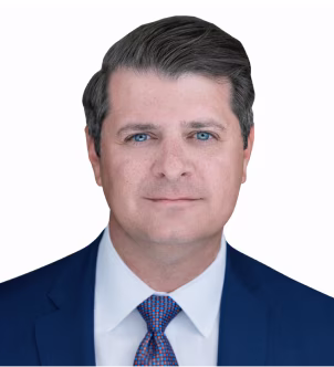Portrait of a man with blue eyes, dark hair, wearing a blue suit, white shirt, and patterned tie against a white background.