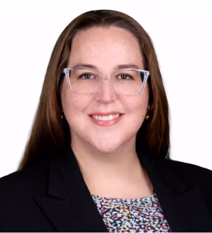 Smiling woman with long dark hair wearing a black blazer and pearl necklace on a white background.