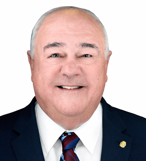 Smiling older man in a dark suit, white shirt, and striped tie with a pin on his lapel, against a white background.