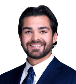A young man with dark hair and beard smiling, dressed in a white shirt, dark blue suit jacket, and dark patterned tie.
