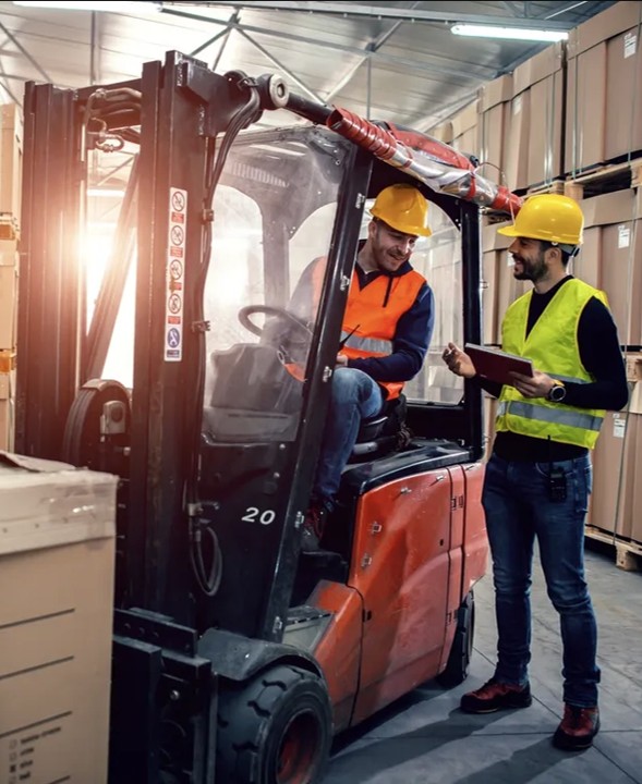 Dos trabajadores con cascos y chalecos de seguridad conversan junto a un montacargas en un almacén con cajas apiladas.