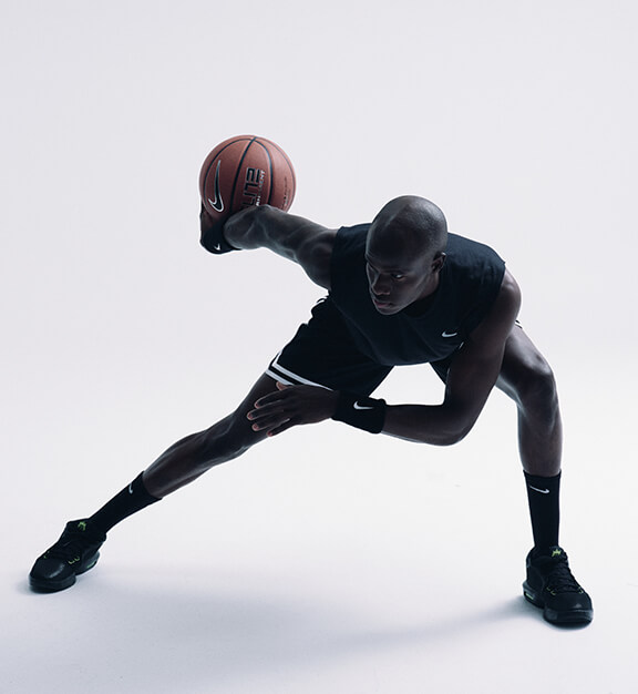 Male basketball player in black athletic wear dribbling a basketball in a wide defensive stance on a plain background.