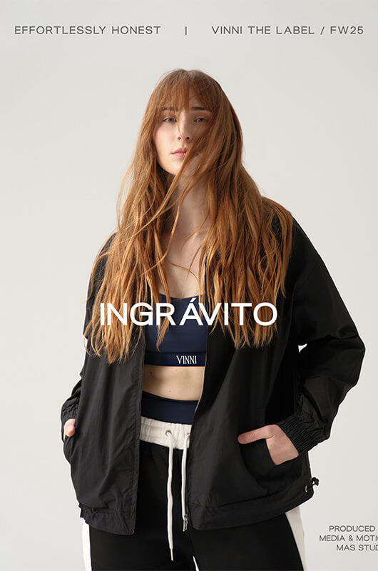 Young woman with long red hair wearing a black jacket over a navy sports bra and black-and-white pants against a plain light background.