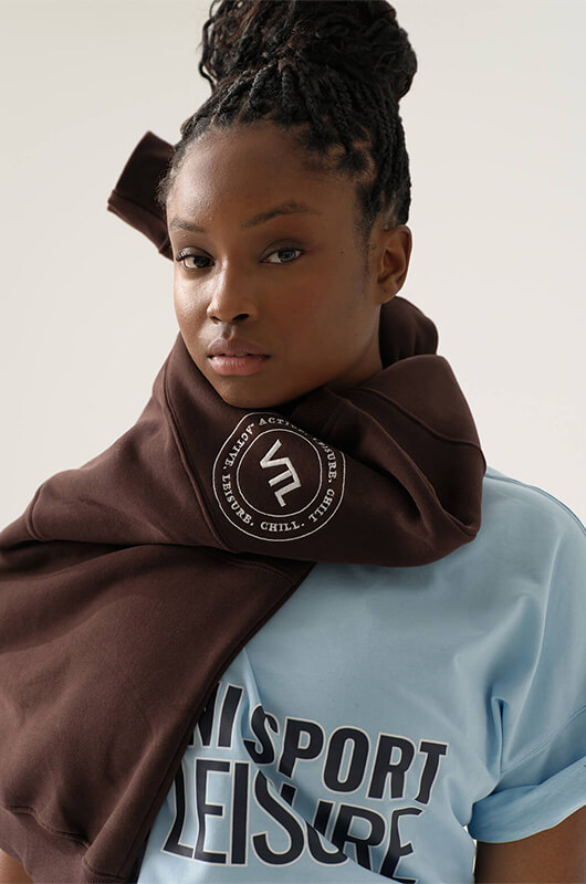 Close-up of a woman wearing a light blue t-shirt with 'UNISPORT LEISURE' text and a brown hoodie draped around her neck with a circular logo patch.