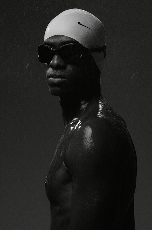 Wet male swimmer wearing white Nike swim cap and goggles looking sideways in black and white photo.
