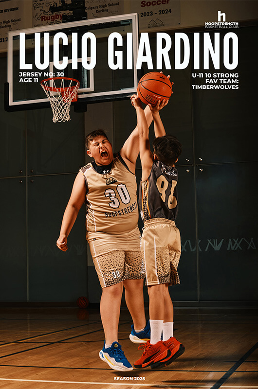 Two young basketball players in beige and black uniforms compete for a rebound under a basketball hoop in a gym.