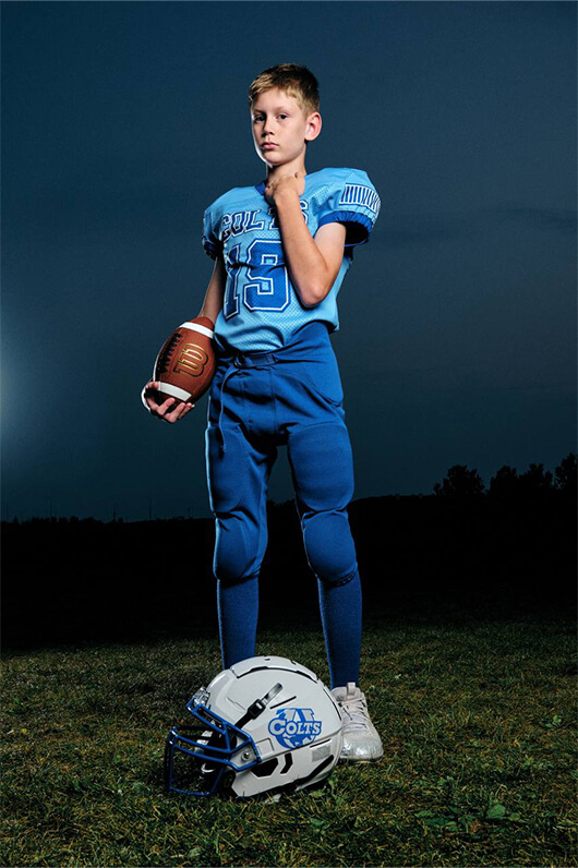 Boy in blue football uniform holding a football with a white Colts helmet on the grass at dusk.