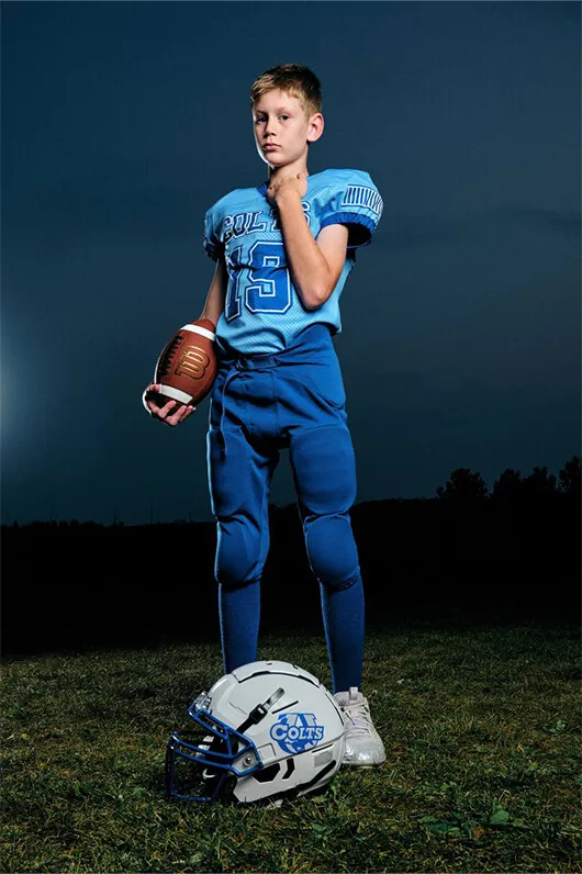 Boy in blue football uniform holding a football with a white Colts helmet on the grass at dusk.