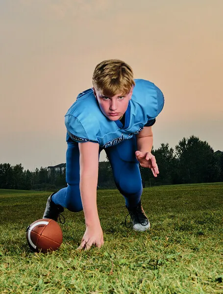 Calgary youth football player in a lineman stance during a Media&Motion outdoor sports media day, captured in full gear at sunset.