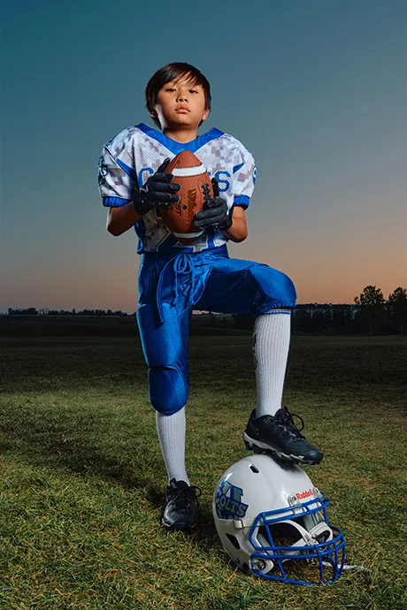 Calgary youth football player from the Colts posing outdoors at sunset during a Media&Motion sports media day, holding a football with helmet at his feet.