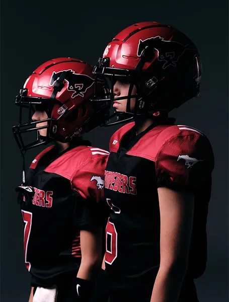 Two youth American football players in red helmets and black red jerseys standing side by side against a dark background.