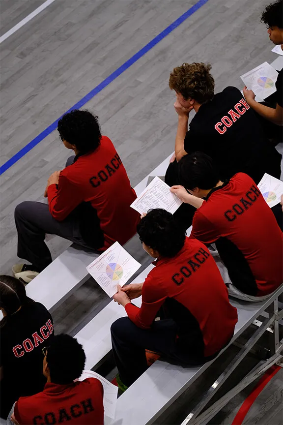 Group of people wearing red and black shirts labeled 'COACH' sitting on bleachers, reviewing papers with colorful charts.