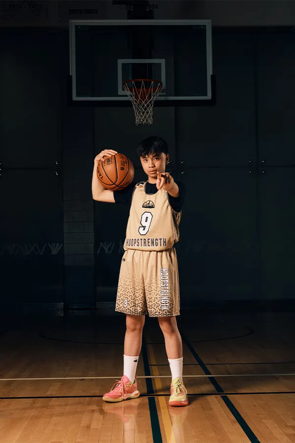 Young basketball player in beige HoopsTrength uniform holding a basketball and pointing forward on indoor court.