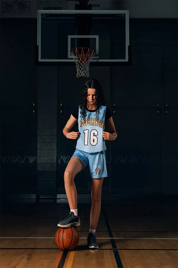 Young female basketball player in blue uniform with number 16 standing in front of a basketball hoop with one foot on a basketball.